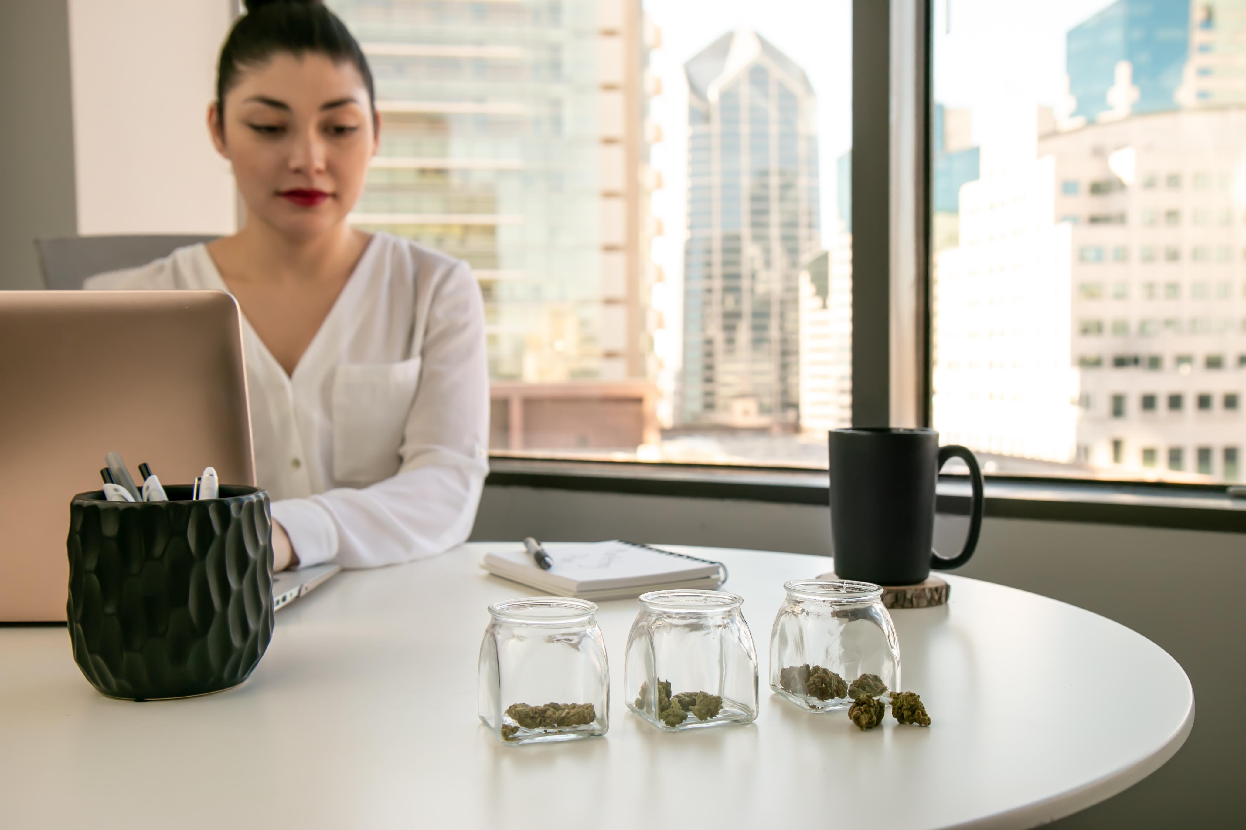 A woman on a laptop looks at three open glass containers of cannabis products placed on her desk