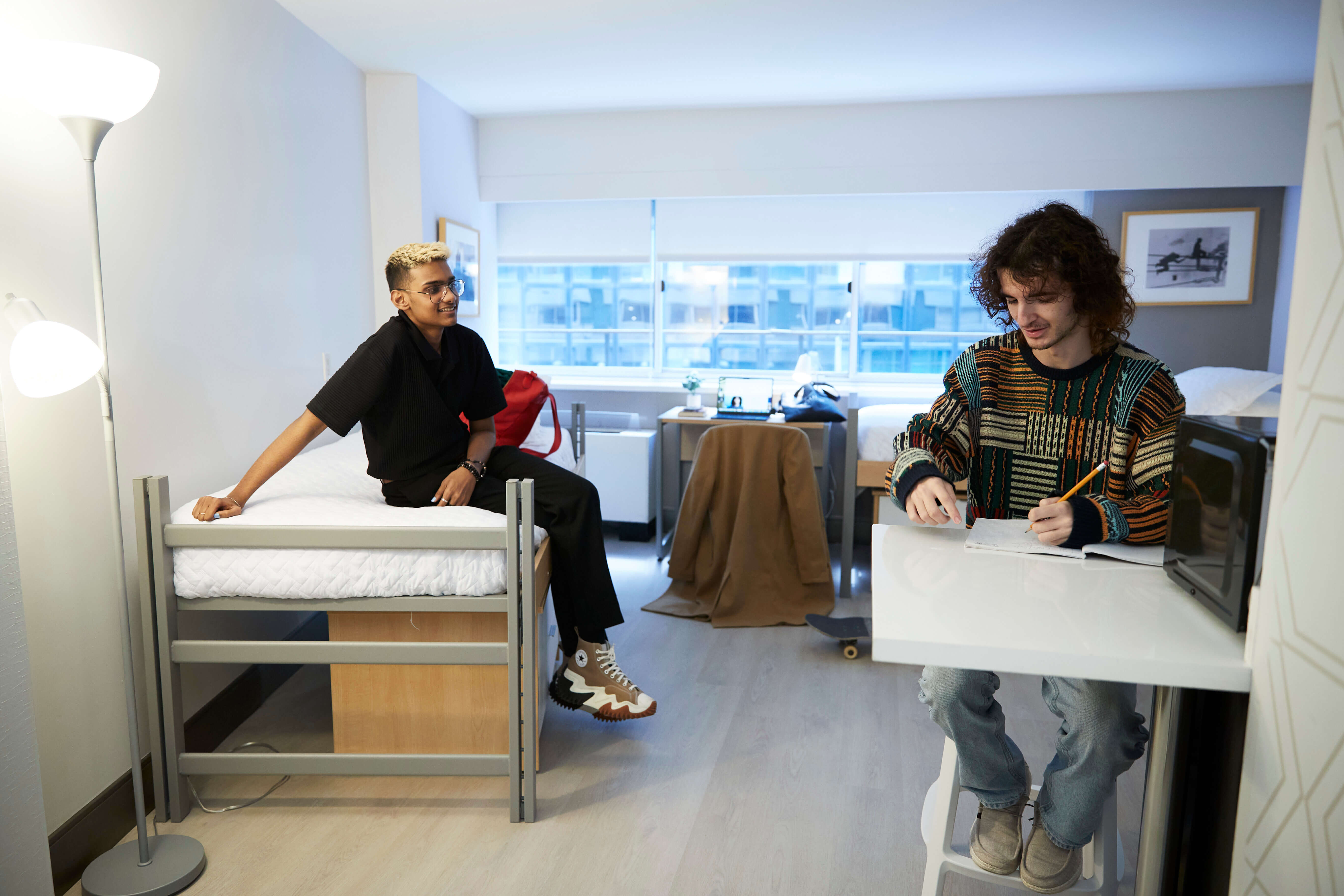 Two LIM students sit in their residence hall room. One is on his bed, while the other sits at a table to sketch