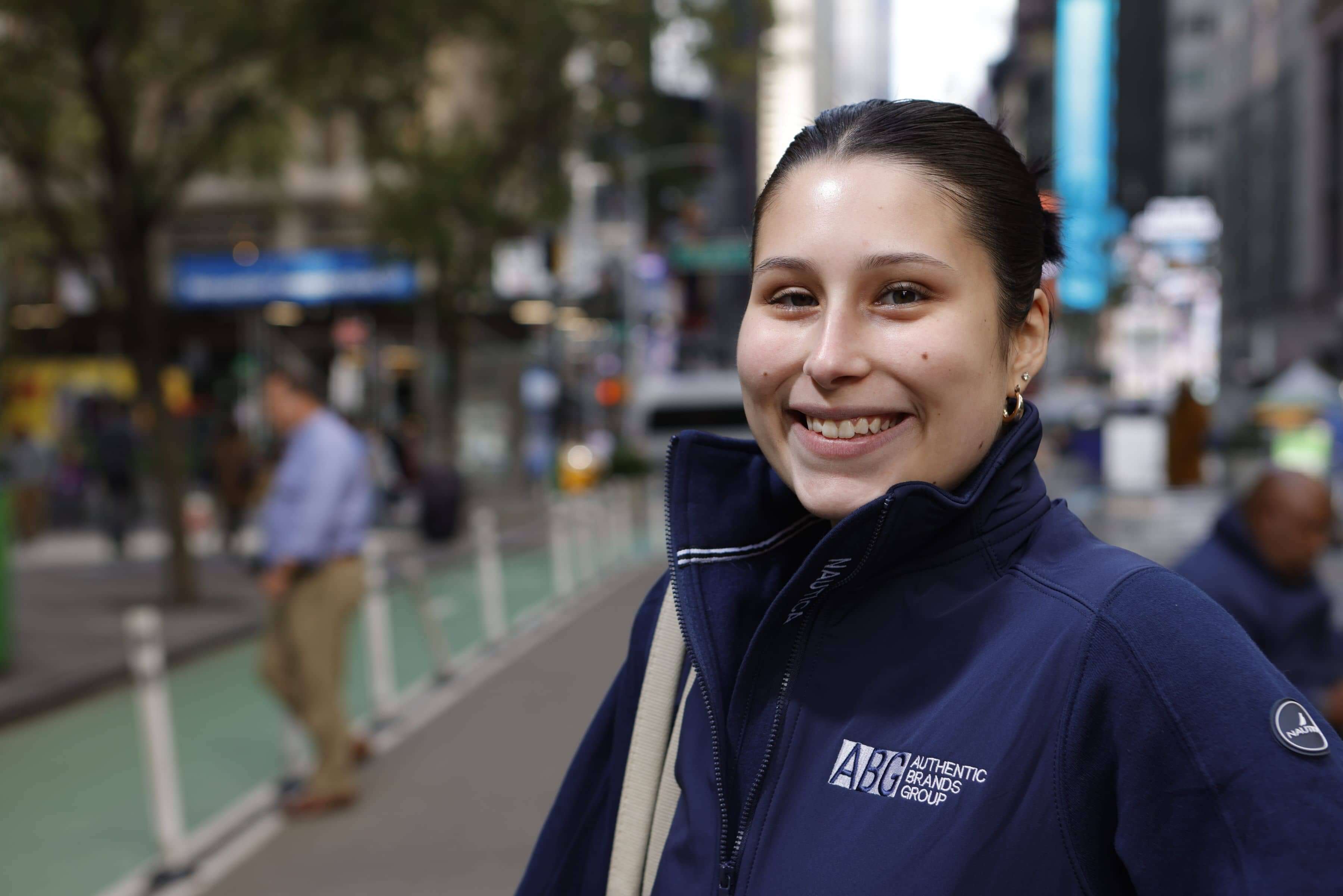 A female LIM student smiles and poses in Midtown. She wears an Authentic Brands Group jacket.