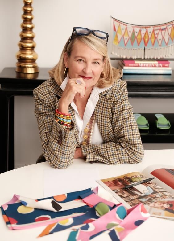 Blonde woman, checkered jacket, glasses, sits at desk with fashion patterns