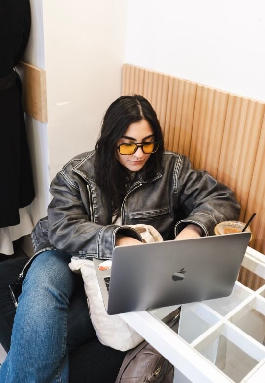 young woman, long dark hair, sunglasses, leather jacket, apple laptop, iced coffee