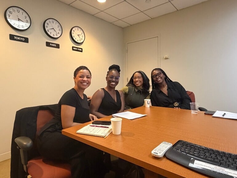 Four women, business casual dress, sit at the end of a conference table on LIM's campus, smiling