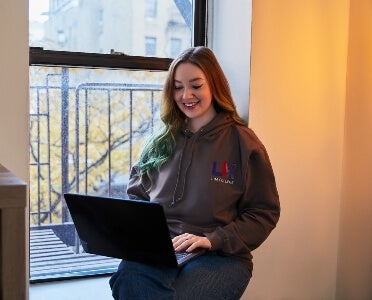 woman, long red hair, LIM College hoodie, sits in a window sill engaged on a laptop