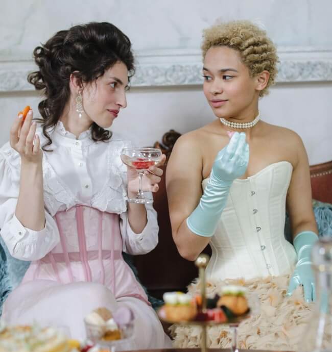 two women in British Regency era attire, eating, looking at one another