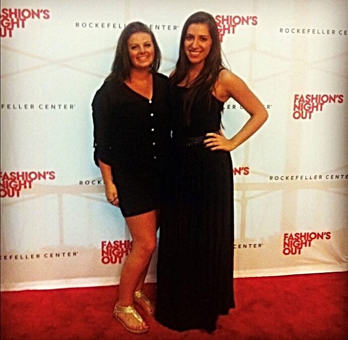 two women, glamorously dressed, stand on a red carpet in front of "Fashion's Night Out" step-and-repeat