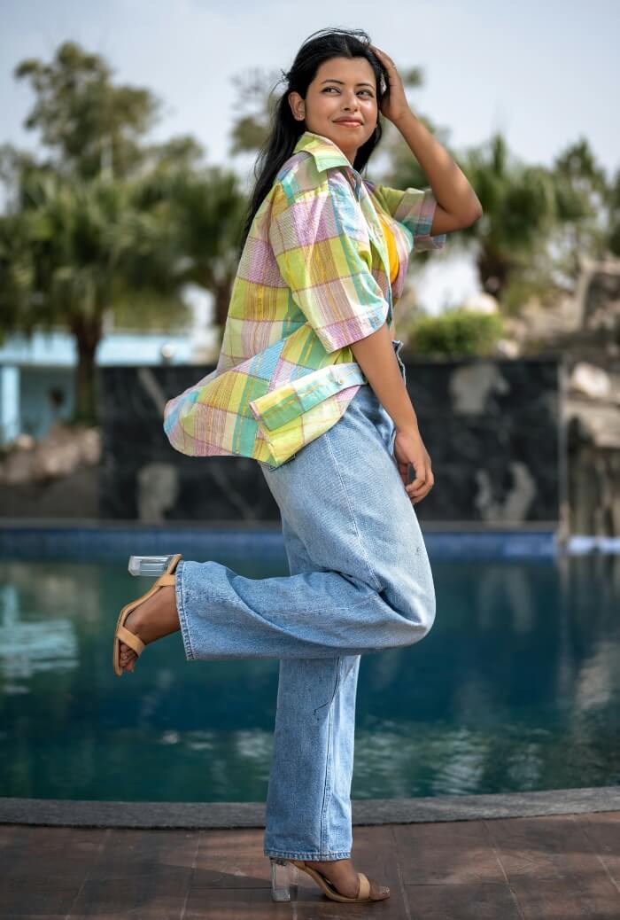 model with long brown hair, brightly patched jacket, jeans, outdoors by water.