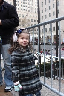 little girl stands at Rockefeller Center in plaid coat and earmuffs