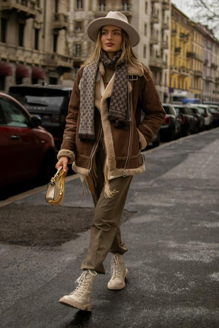 Maximalism example, woman in scarf, thick-trim coat, fedora hat, gold purse, waking down a city street.