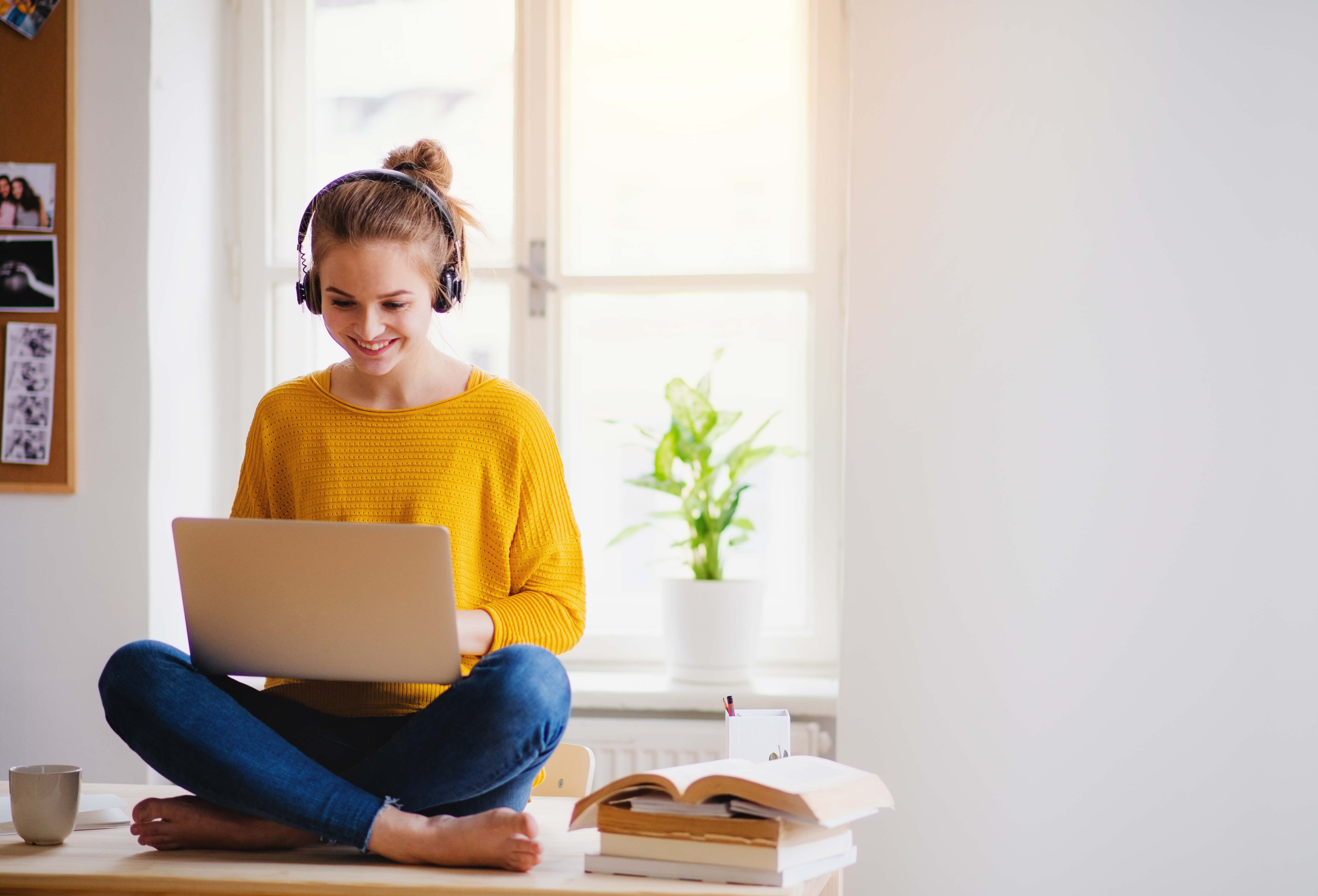 A woman in a yellow sweater and jeans sits on a table top wearing headphones and typing on a laptop on her lap