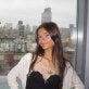 young woman, long brown hair, stands in front of a New York City window