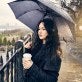 young woman, long black hair, stands on Brooklyn promenade in New York holding coffee and an umbrella