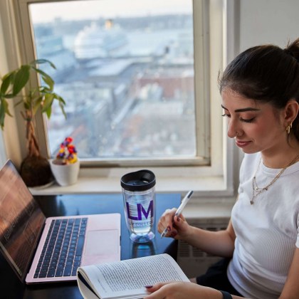 A students sits at a table next to a window with a laptop and book open