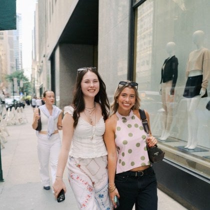 two young female students walk down 5th Avenue in New York City by a fashion business window display.