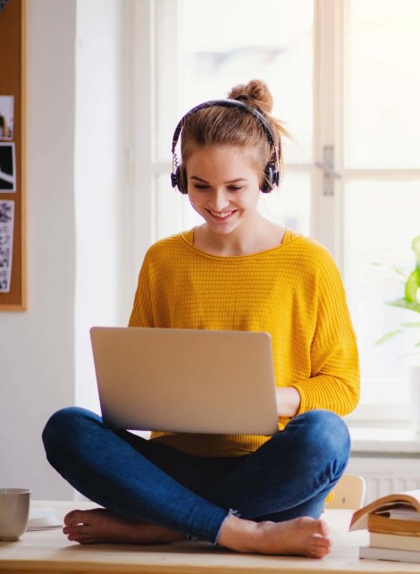 A woman sitting on a tabletop has a laptop on her lap. She is wearing headphones and a yellow shirt. 