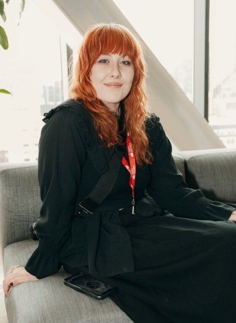 young woman, long red hair, black coat and dress, seated on a gray couch