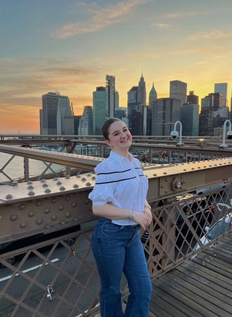 young woman, jeans and white shirt, stands on Brooklyn Bridge with New York City skyline in the background