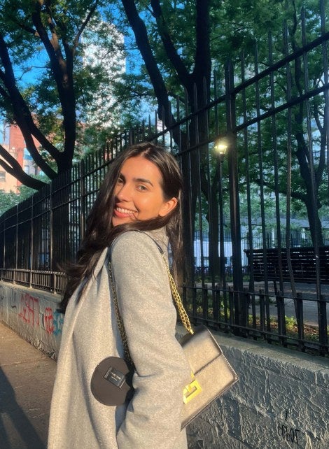 young woman, long dark hair, standing near iron fence in New York City, wearing light-colored coat