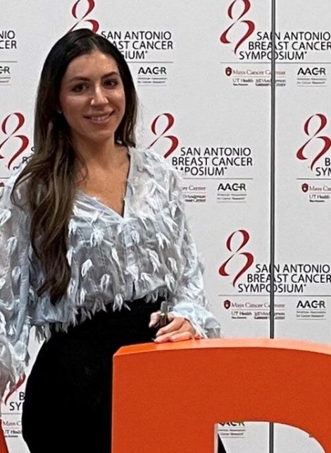 woman, long dark hair, stands in front of breast cancer awareness step-and-repeat