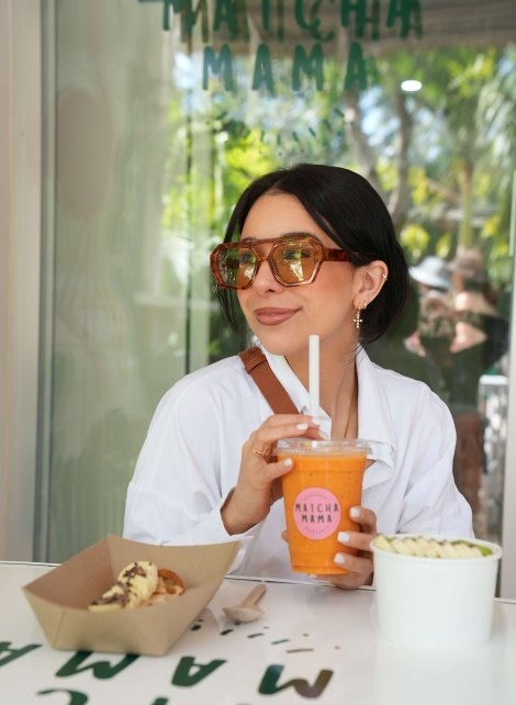 young woman, dark hair, sunglasses, white shirt, drinks an orange Matcha Mama drink 