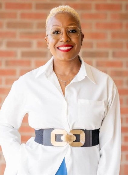 Woman, short hair, glasses, white professional shirt, stands against brick wall.