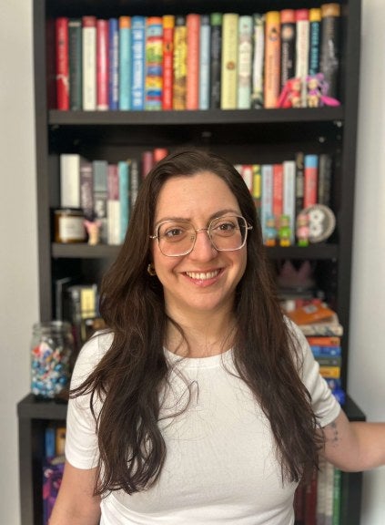 woman, long dark hair, glasses, white shirt, stands in front of bookcase