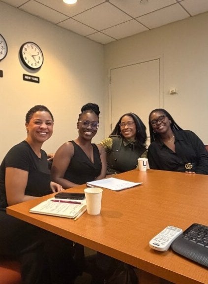 Four women, business casual dress, sit at the end of a conference table on LIM's campus, smiling
