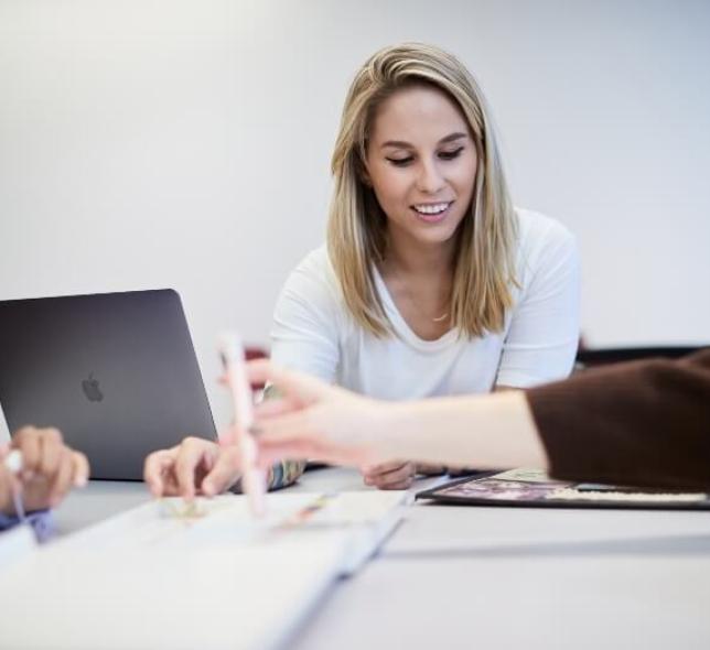 blonde female student, white t-shirt, in open concept classroom with laptop