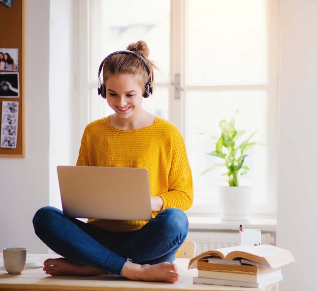 A woman sitting on a tabletop has a laptop on her lap. She is wearing headphones and a yellow shirt. 