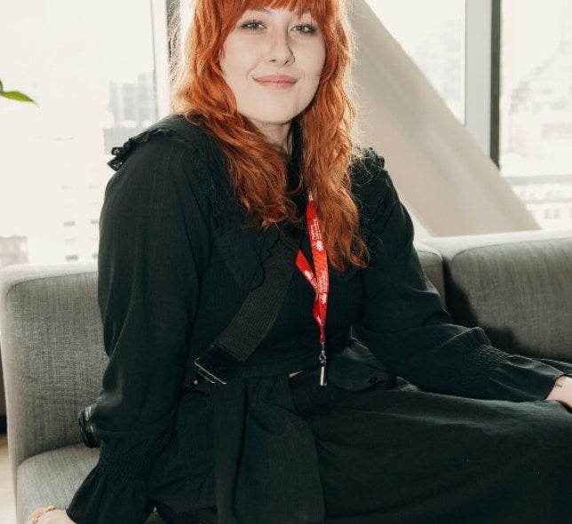 young woman, long red hair, black coat and dress, seated on a gray couch