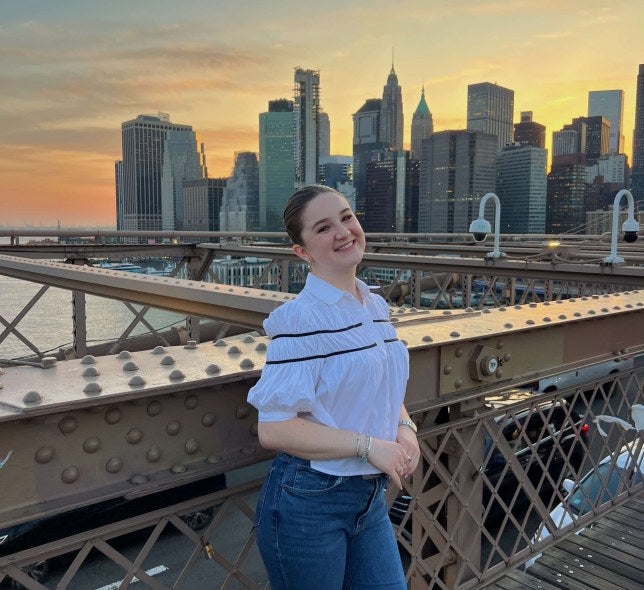 young woman, jeans and white shirt, stands on Brooklyn Bridge with New York City skyline in the background