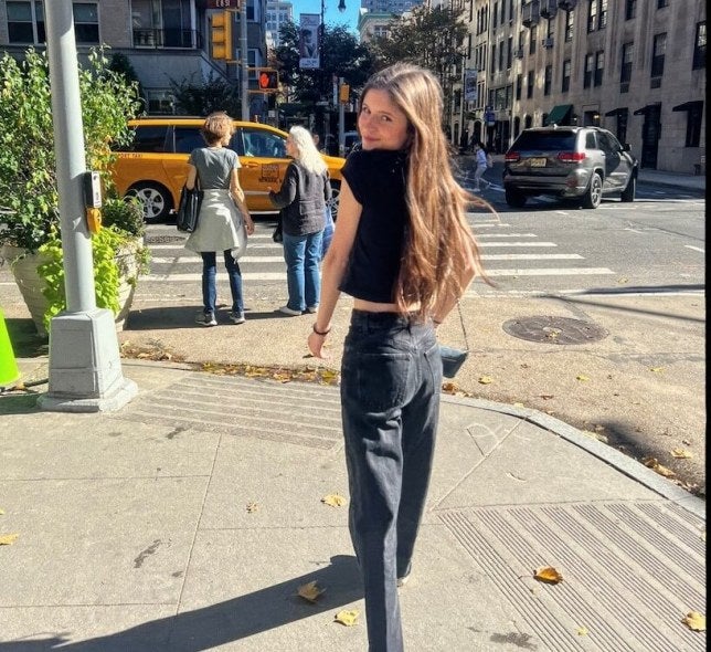 young woman, long brown hair, jeans, dark shirt, New York City street corner