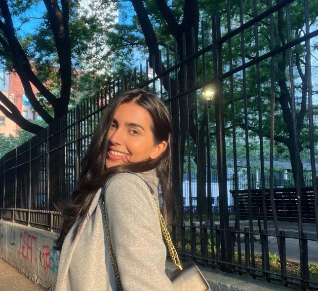 young woman, long dark hair, standing near iron fence in New York City, wearing light-colored coat
