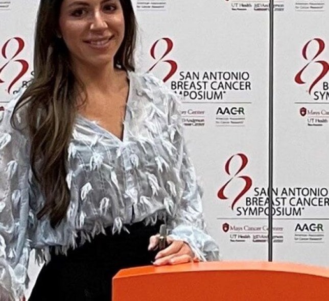 woman, long dark hair, stands in front of breast cancer awareness step-and-repeat