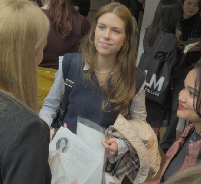 woman handing resume to a recruiter at a busy career fair event, an LIM College tote bag is in the background.