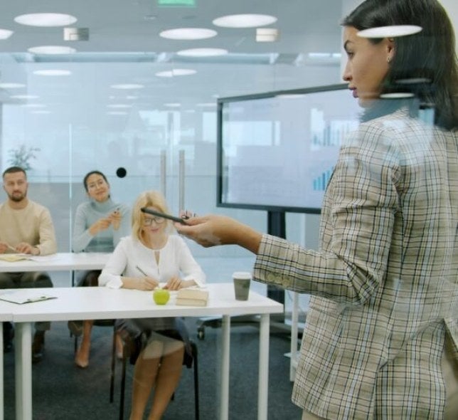young woman, brown hair, patterned jacket, presents in a meeting room in a corporate setting.