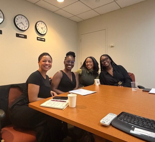 Four women, business casual dress, sit at the end of a conference table on LIM's campus, smiling
