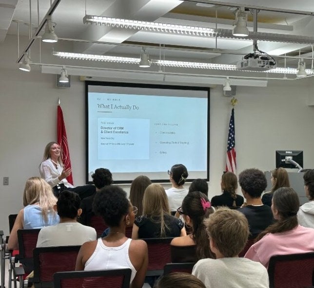 guest speaker, long hair, white shirt, in front of event space filled with student spectators