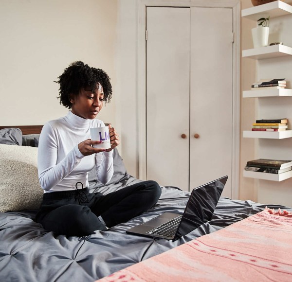 A student sits on a bed holding a coffee cup looking at a laptop