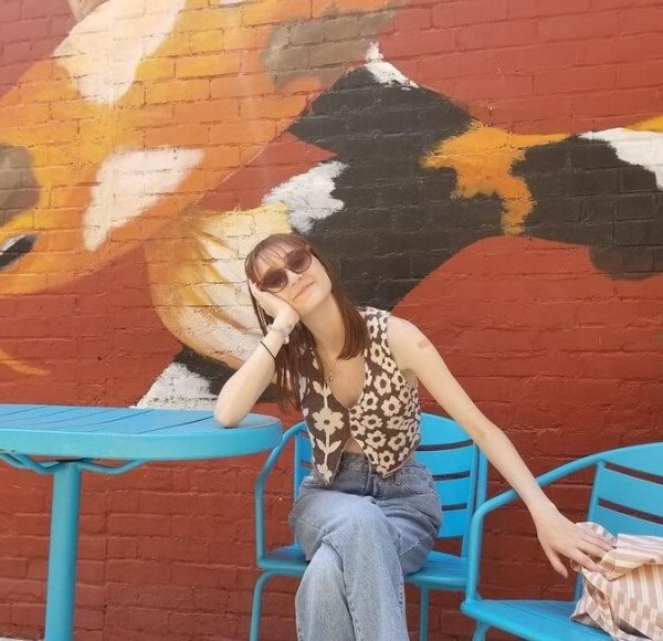 Ella Sutton sitting in a blue chair, leaning on a table in front of a mural of koi fish on a brick wall