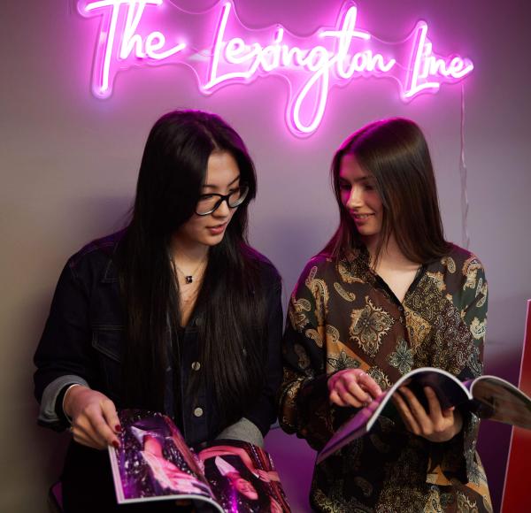 Two LIM students look through the latest issue of The Lexington Line under a neon sign that reads The Lexington Line