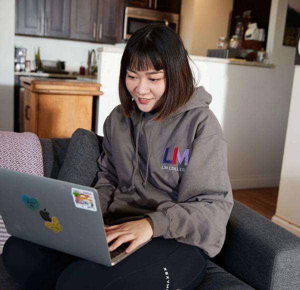 An LIM student wearing an LIM hoodie goes on her laptop in the living room of an apartment