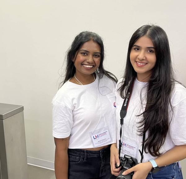 Two LIM students smile and pose for the camera. Both wear white t-shirts and one holds a DSLR camera
