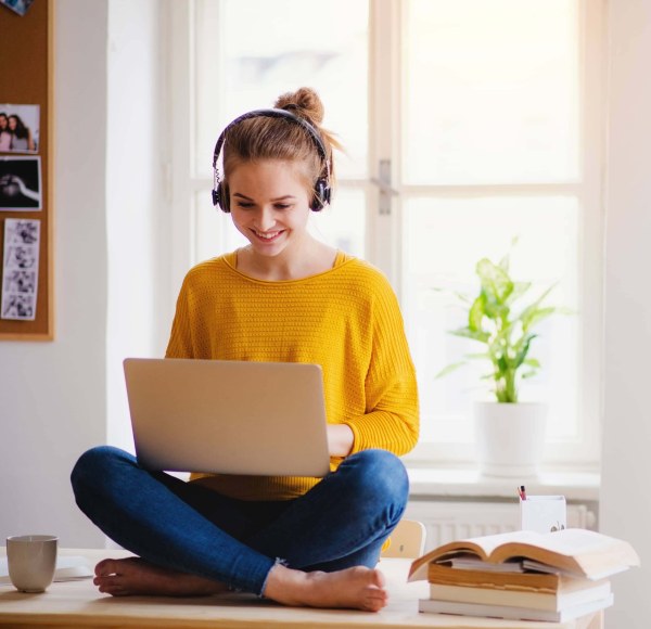 A woman sitting on a tabletop has a laptop on her lap. She is wearing headphones and a yellow shirt. 