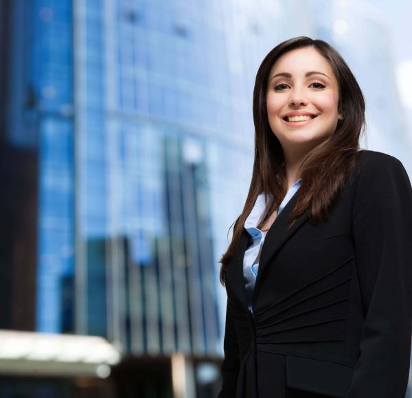 A woman in business attire smiles with tall glass buildings in the background