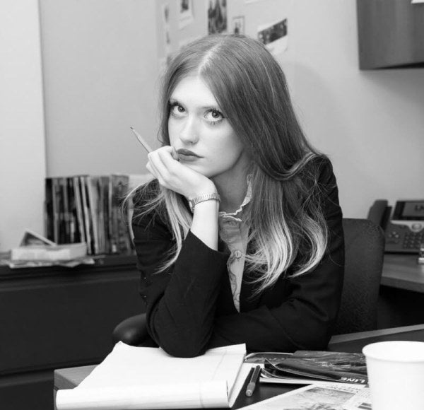 Black and white photo, young woman, long red hair professionally dressed, sitting at desk.