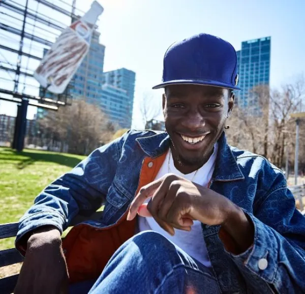 young man, blue hat, denim jacket, jeans sits on bench in Queensbridge Park.