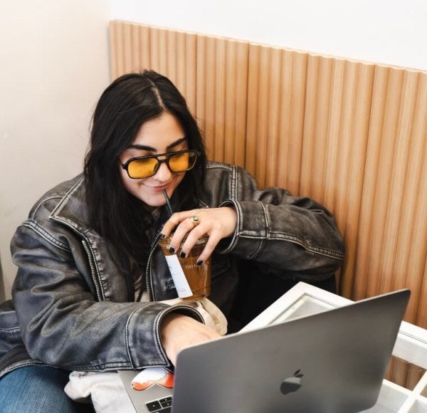 young woman, long dark hair, leather jacket, laptop, tinted sunglasses, sipping iced coffee