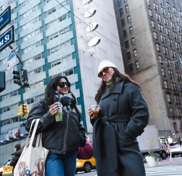 two students stand outside the FOUND Study - Midtown East residence hall in winter, wearing sunglasses, drinking coffee