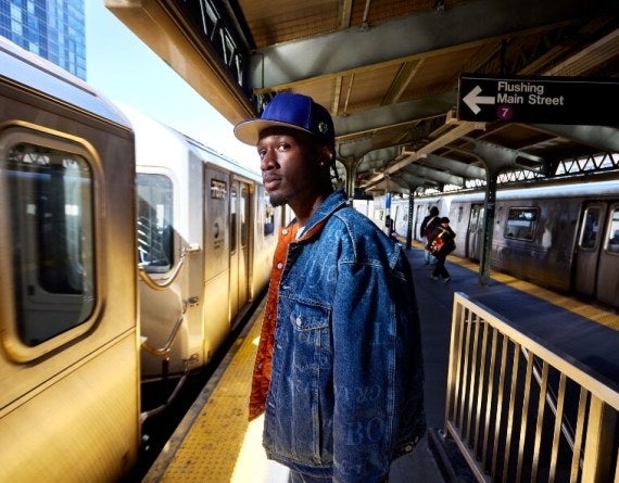 A male student in a denim jacket and blue hat waits on the 7 train platform in Queens, New York.