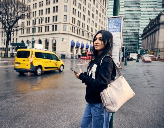young woman, long dark hair, LIM College sweatshirt, waits at a bus stop while using her smartphone